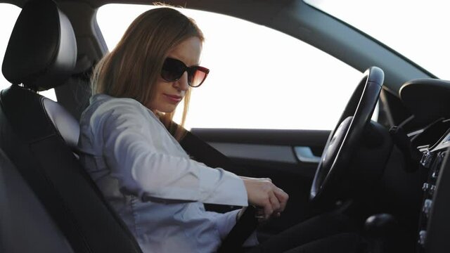 Side view of mature caucasian woman in sunglasses and formal wear fastening seatbelt before driving car. Safety driving and responsibility ly concept.
