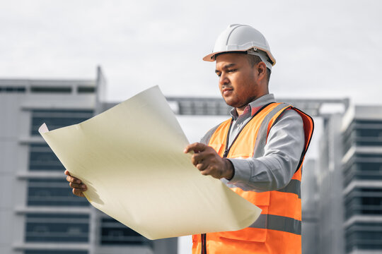 Asian engineer handsome man or architect open the paperwork blueprint and planning to building construction. Worker asian man with white safety helmet working in modern building construction site.