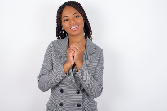 Dreamy charming Young African American businesswoman with braids on white wall with pleasant expression, keeps hands crossed near face, excited about something pleasant.