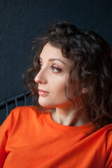 Fototapeta premium Female portrait of a beautiful curly girl in bright orange t-shirt at home in her apartment on dark background, happy people concept
