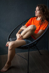 Obraz na płótnie Canvas Female portrait of a beautiful curly girl in bright orange t-shirt sitting at home in her apartment on dark background, happy people concept