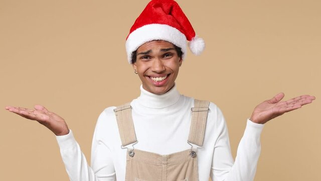 Confused young Santa curly african american man 20s in white shirt Christmas hat look camera spread hands say oops ouch oh omg i am so sorry isolated on plain pastel beige background studio portrait
