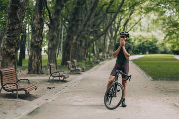 Fototapeta na wymiar Tired sportsman standing at green summer park with bike