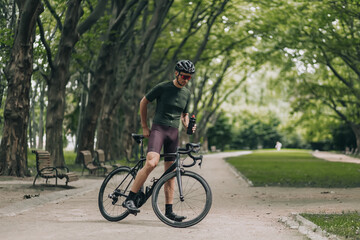 Fototapeta na wymiar Cyclist in helmet and glasses drinking water at park
