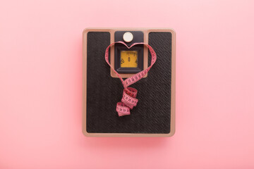 Heart shape created from rolled out measure tape on weight scales on light pink floor background. Pastel color. Closeup. Female body weighing and measuring concept. Top down view.