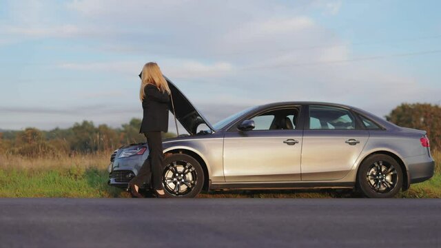 Mature business woman in eyeglasses and black suit standing near broken car with open hood and talking on mobile. Female driver calling someone for help during problems on road.