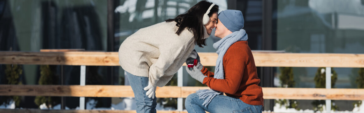 happy man holding box with wedding ring and kneeling while making proposal to woman on ice rink, banner