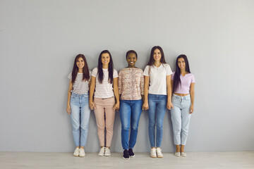 Portrait of beautiful smiling young diverse women standing in row holding hands. Multiracial women in casual clothes look at the camera on a background of a light wall. Women's diversity concept.