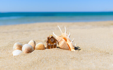 A small Christmas toy in the shape of a gingerbread house stands on a sandy beach next to beautiful shells in the background of the sea. Selective focus on the house.
