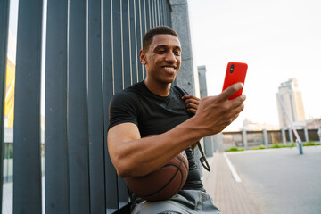 Black sportsman using cellphone while sitting with basketball