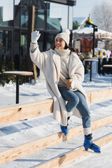 full length of happy young woman in winter hat and ice skates holding paper cup and waving hand near ice rink