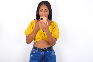afro american woman with braids wearing sexy yellow t-shirt on white wall being deeply surprised,...