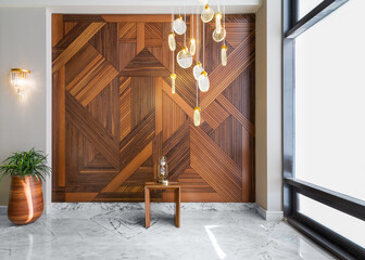 Hall with wood cladding wall decorated with geometrical patterns, tall rounded wooden planter with green bushes, contemporary glass tall chandelier, big window, and white marble floor in modern house