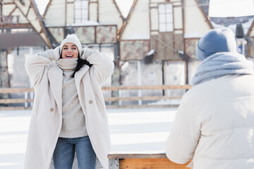 smiling young woman adjusting winter hat near blurred boyfriend