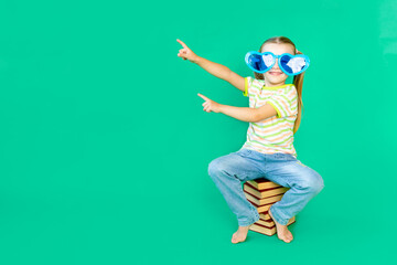 Fototapeta na wymiar a girl child sits on a green background on books in blue large funny glasses and points her finger up or to the side, space for text