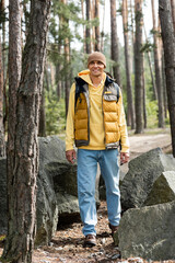 full length view of smiling tourist in warm vest and beanie looking away in forest