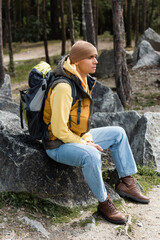 traveler with backpack, wearing warm vest and beanie, sitting on stone in forest