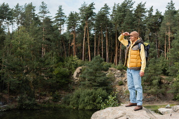 Naklejka na ściany i meble full length view of tourist standing on rocky cliff near forest lake and looking away