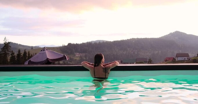 Rear view of beautiful tattooed woman in swimsuit leaning at the edge of pool admiring scenic mountain forest