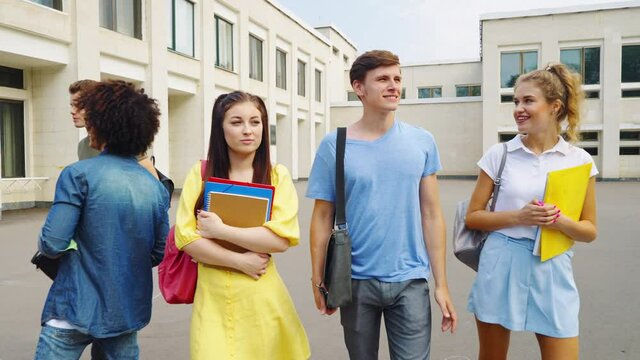 Students leaving college after classes, shy female hugging books with both hands, another female looking with admiration at man in the center, group of students standing nearby. Concept of relations