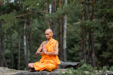 buddhist monk in orange kasaya sitting in lotus pose with praying hands while meditating in forest