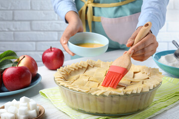 Woman spreading egg yolk onto raw apple pie at white wooden table, closeup