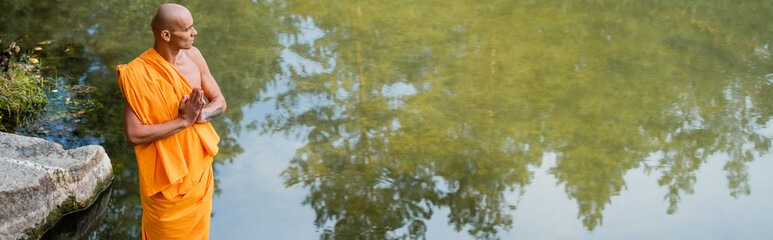 high angle view of buddhist monk looking at lake while meditating with praying hands, banner