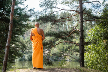 back view of buddhist in orange kasaya meditating in forest near lake