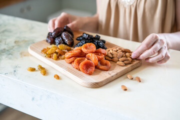 preparing a healthy breakfast, dried apricots, raisin, prunes, dates and almonds on a wooden cutting board in the kitchen on the table. Dried fruit in female hands