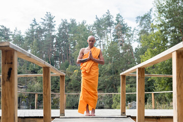 full length view of buddhist monk praying on wooden platform in forest