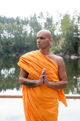 buddhist monk in traditional robe looking away while meditating near lake in forest