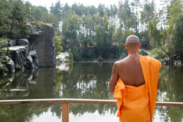 back view of buddhist in orange kasaya meditating near forest lake
