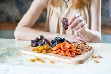 preparing a healthy breakfast, dried apricots, raisin, prunes, dates and almonds on a wooden cutting board in the kitchen on the table. Dried fruit in female hands