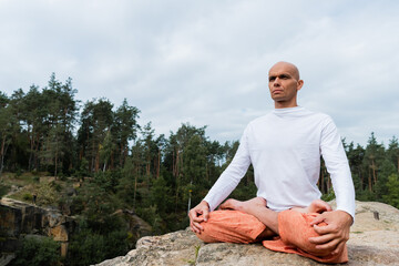 Fototapeta na wymiar buddhist in sweatshirt looking away while meditating in lotus pose outdoors