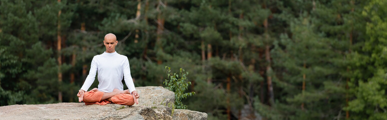 buddhist in white sweatshirt practicing lotus pose on rock in forest, banner