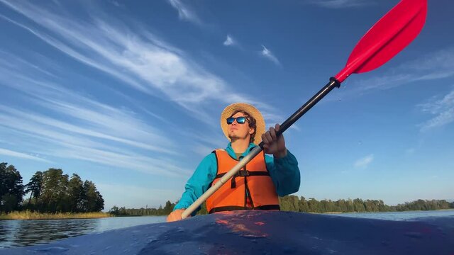 Young man in straw hat sunglasses and orange life jacket rows paddle sitting in boat against river bank forest under blue sky closeup