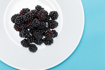 A white plate with blackberries on a blue background. Useful forest fruits. Super food. Concept of healthy eating. top view. Close-up. Macro. Blackberry berries with water drops. Flat lay