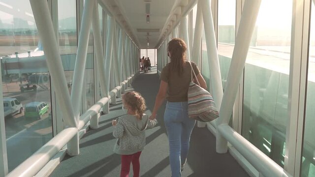 Back view: A young woman holding her daughter's hand is walking the telescopic gangway, sleeve, aerobridge in plane in airport for boarding on aircraft. Traveling with children, flying on vacation.
