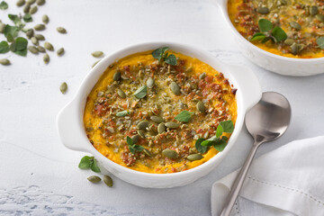 Homemade pumpkin casserole or gratin with cheese and pumpkin seeds in two ceramic tins on a light gray background