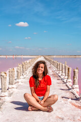 Fototapeta na wymiar A cute young woman is sitting on a salty beach between wooden sticks on a salty pink lake with a blue sky. A peaceful landscape and relaxation