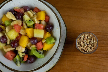 Refreshing fruit salad, natural colors, textures and different cuts, served in a crockery container and a small white plate, with natural cereals served in a transparent container on the side, wooden 