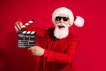 Photo of funny grey hair elder man hold clap wear santa cap red suit eyewear isolated on red color...