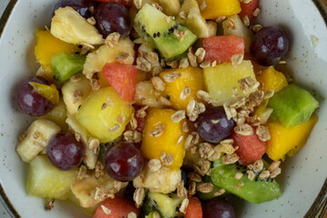 Refreshing fruit salad with natural cereals served on white dinnerware, wooden background, close up, top view