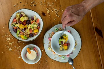 man serving fruit salad with silver spoon, in white bowl, decorated with colorful cereals, and yogurt on wooden table in photo taken from above.