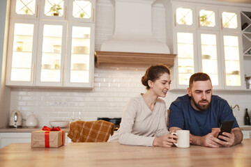 Cheerful young Caucasian couple sitting at table in kitchen and drinking coffee while surfing net together on smartphone
