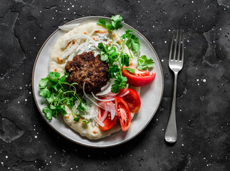 Unleavened flatbread with burger, yogurt sauce, fresh tomatoes and cilantro on a dark background,...