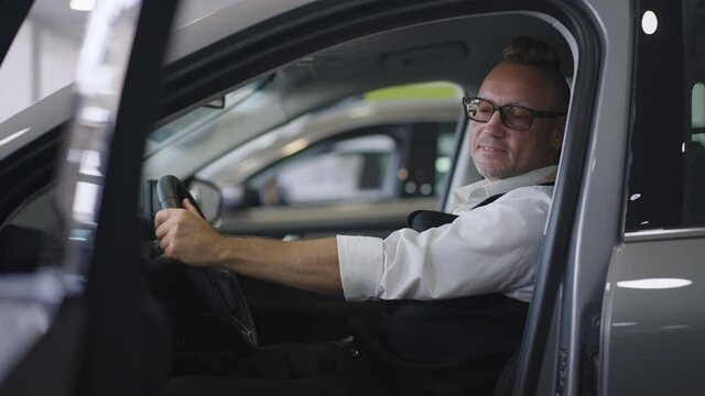 Side view portrait of confident successful Caucasian man in eyeglasses with pierced ear sitting in vehicle examining interiors. Wealthy customer choosing automobile in dealership indoors. Slow motion