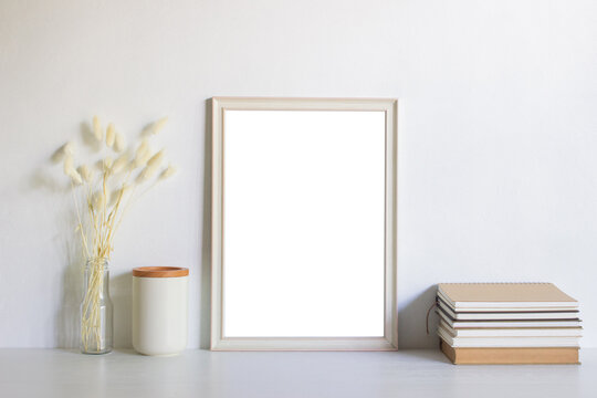 A white blank photo frame with books and flowers over the white wall. 