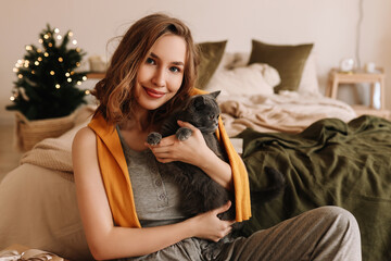 A pretty smiling young woman in pajamas hugs her friend's pet domestic cat sitting in a cozy bedroom decorated with Christmas decor at home, selective focus