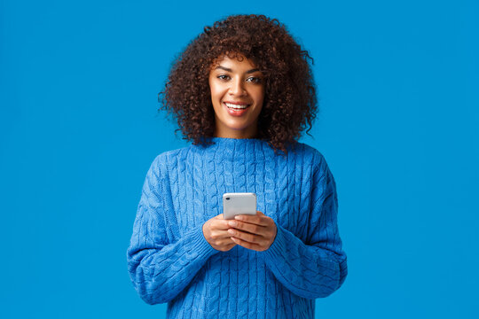 Smartphone addication, gen-z and people concept. Charismatic lovely smiling african-american woman with afro haircut in winter sweater, holding smartphone and looking camera, blue background